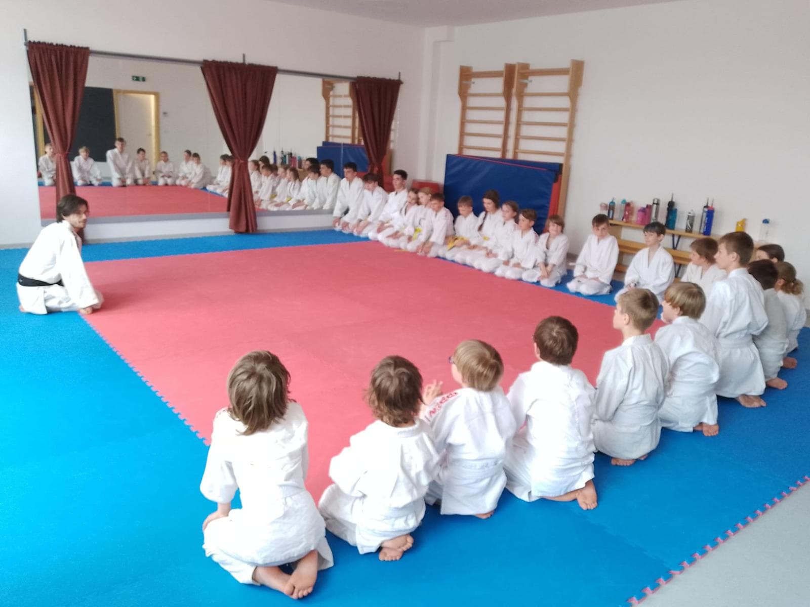 Trenér klečí před skupinou dětí sedících v kruhu na začátku tréninku